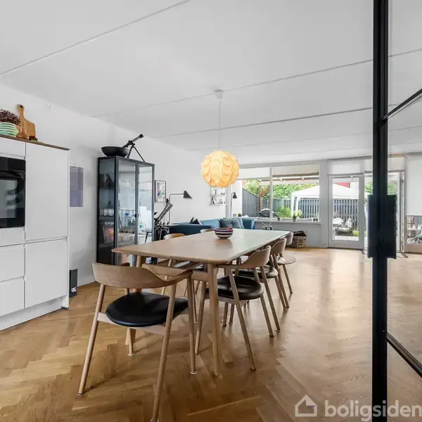 Et spisebord med træstole står i en moderne, lyse stue. En stor glasdør adskiller spiseområdet fra opholdsafdelingen. Udsigt til terrassen gennem store vinduer.