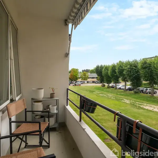 Balkon med to træstole og et lille bord. Udsigt til græsplæne og parkeringsplads med træer i baggrunden. Klar blå himmel og justerbar markise over balkon.