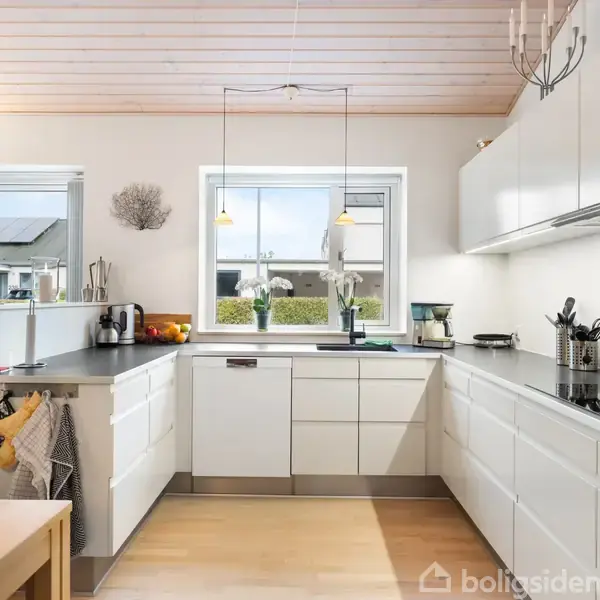 Et moderne køkken med hvide skabe og sort bordplade. En vaskemodul står foran et stort vindue. Spisebord til venstre. Lyse trælofter og gulve.