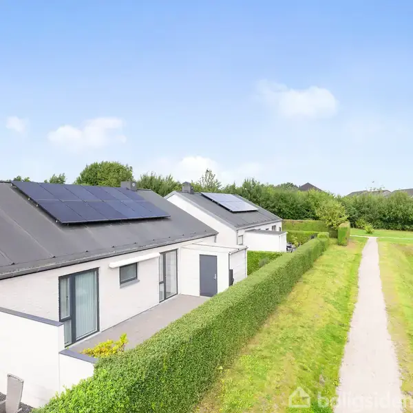 Hus med solpaneler på taget står langs en grusvej omgivet af hække og græsarealer. I baggrunden ses buske og træer mod en klar blå himmel.