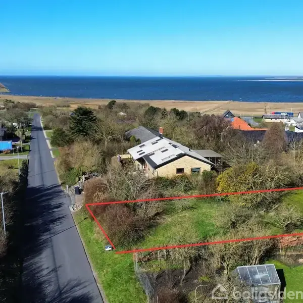 En rektangulær, græsbevokset grund er markeret med rødt. Den er omgivet af huse og træer. I baggrunden ses en vej og havet.