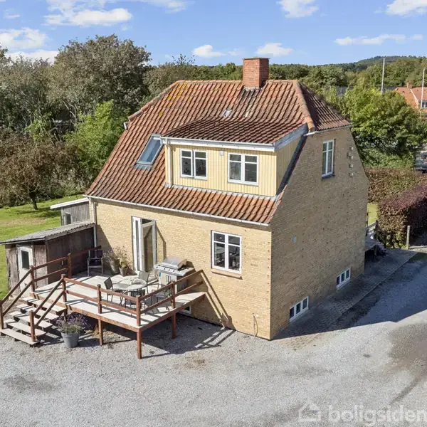 Et to-etagers hus med rødt tegltag står i en have med græs og omgivende træer. Foran huset er en terrasse med borde og stole.