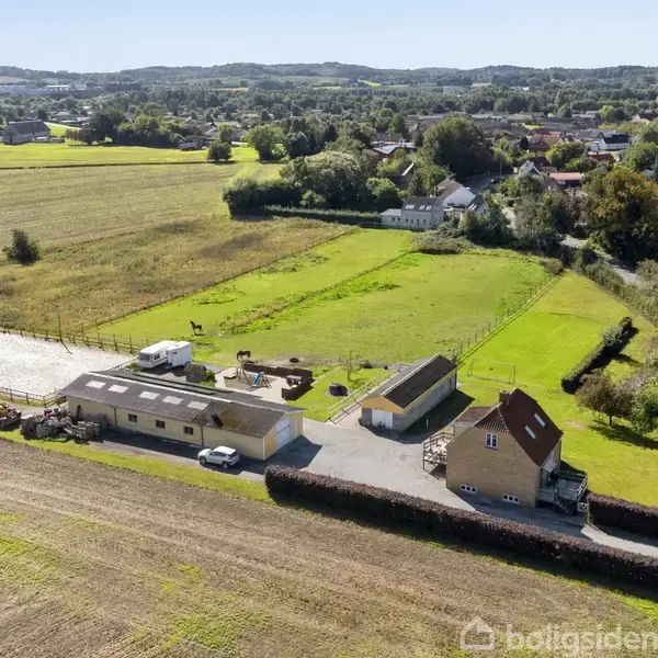 En gård med stalde og beboelseshus ligger ved siden af grønne marker og en lille landsby med træer og huse i baggrunden.