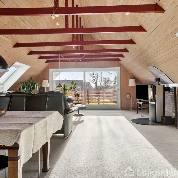 Stue med spisebord og lædersofa arrangeret under en loft af træbjælker. Sollys strømmer ind gennem skråvinduer, mens en skydedør fører til altan. Pejs og TV ved væggen.