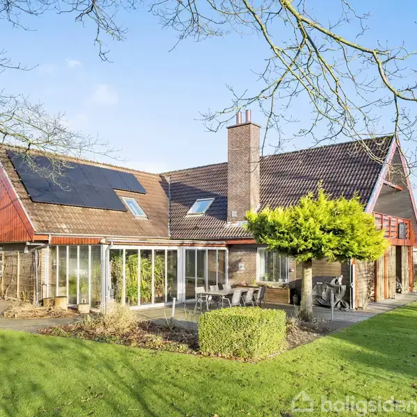 Hus med store vinduer står stille i en velplejet have med grøn græsplæne, buske og træer. Solpaneler er installeret på taget.
