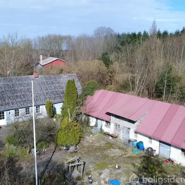 Et landligt hus og en lade med rødt tag står stille omgivet af træer og buske. En grusbelagt gårdsplads ligger imellem dem, indrammet af natur.