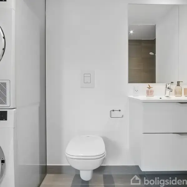 Vaskemaskine og tørretumbler i søjle står i moderne badeværelse med væghængt toilet og hvid vask på skab med spejl. Indretning er minimalistisk og lys.
