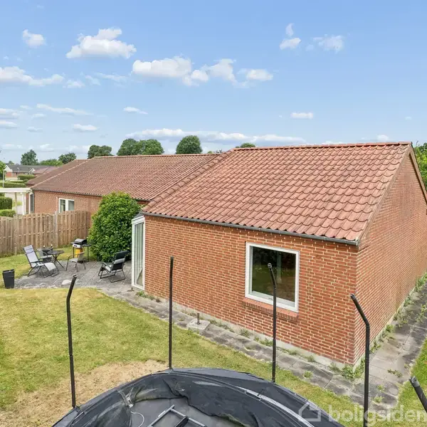 Et rødt murstenshus med tagsten står i en grøn have. En trampolin er placeret foran huset. Haven har havemøbler og er omgivet af en hæk.