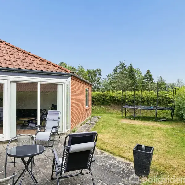 Rødt murstenshus med store vinduer. Terrasse med havemøbler i forgrunden. Trampolin på en græsplæne bagved, omgivet af hække og træer. Blå himmel indikerer solskinsvejr.