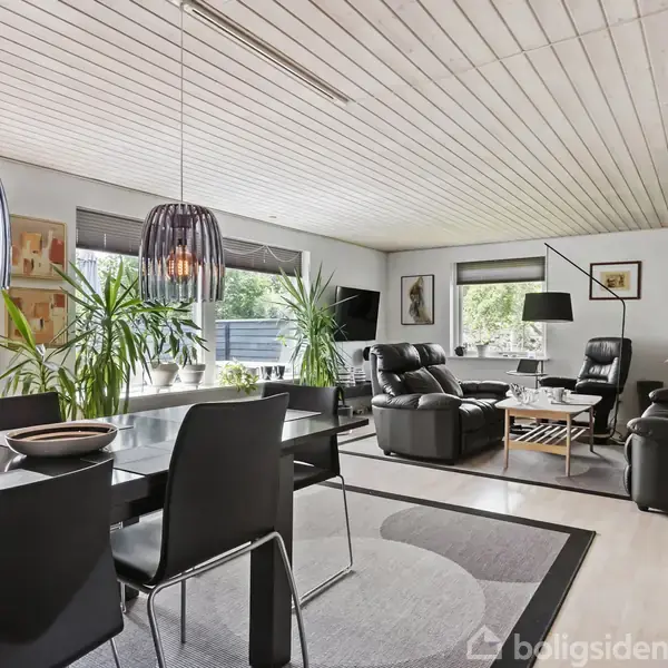 En lys stue med sort lædersofa, stående ved et sofabord. Store vinduer og indendørs planter omkranser rummet. To moderne lamper hænger over et spisebord med sorte stole.