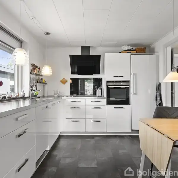 Hvidt køkken med moderne skabe og apparater. En indbygget ovn og kogeplade er centralt placeret. Et bord med stole er i forgrunden, og vinduer giver lysindfald fra venstre.