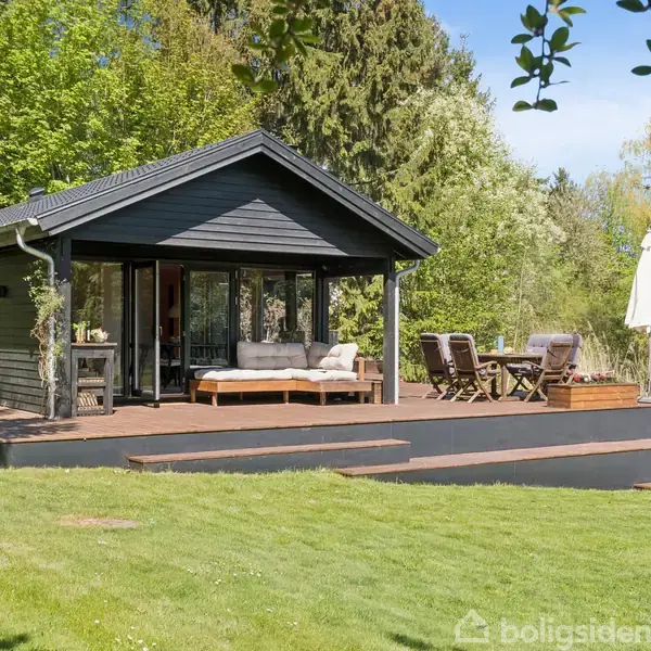 Sommerhus med sort træfacade står stille på en træterrasse med sofa og spisebordssæt, omgivet af frodig grøn vegetation og græsplæne, under en lysblå, klar himmel.