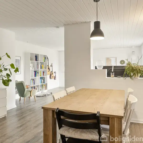 Spisebord med stole står i et lyst rum. En hængelampe hænger over bordet. I baggrunden ses en reol, grønne planter og moderne møbler.