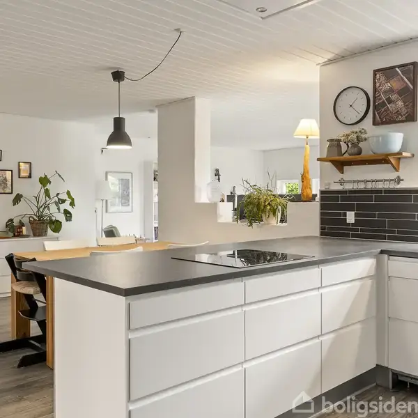 Hvidt køkken med moderne skabs- og skuffeindretning; kogeplader integreret i bordpladen. Mørke fliser pryder væggen over bordet. Stue i baggrunden har spisebord, planter og vægdekorationer.