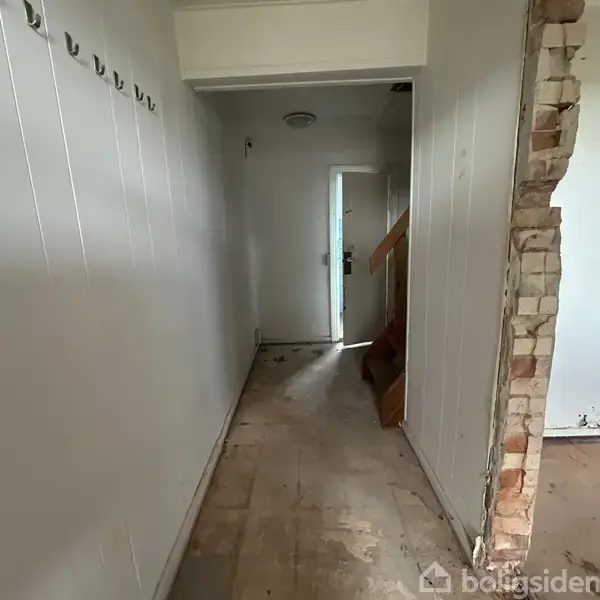 En smal gang fører til en lukket dør. Gulvet er slidt, og en døråbning til højre viser delvist synlige mursten. Væggen til venstre har kroge.