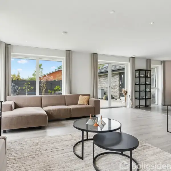 En moderne stue har to store, lyse sofaer og runde sofaborde på et tæppe. Store vinduer lader lys trænge ind, og et spisebord er synligt i baggrunden.