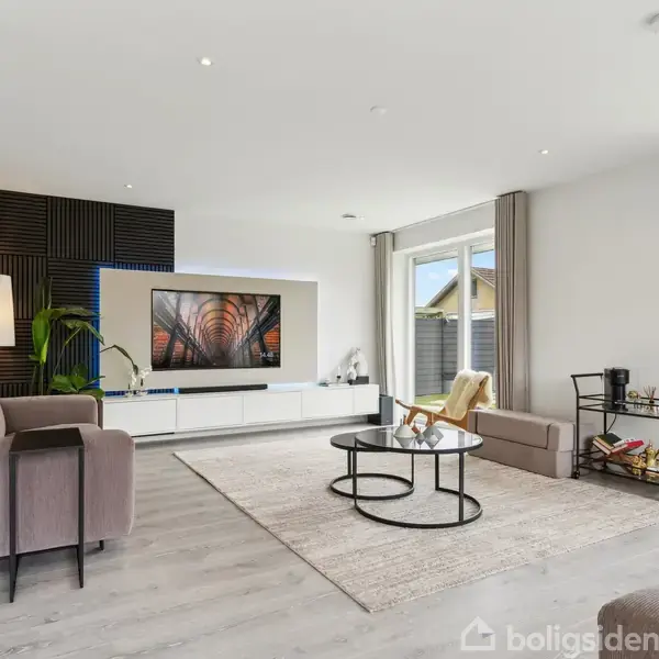 To sofaer og sofabord står i en stue med moderne indretning. En stor plante og en lampe er placeret i hjørnet. På væggen hænger et fladskærms-tv. Store vinduer giver udsigt til en have.