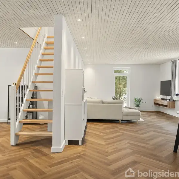 En åben stue med en trappe til venstre fører opad. Sofaen står foran et vindue, og et spisebord er til højre. Parketgulve gennem hele rummet.