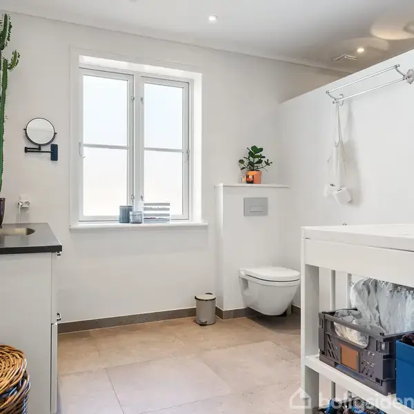 Badeværelset har et toilet monteret på væggen, en hvid vask med planter på bordet og kurve på gulvet, oplyst af et stort vindue med neutrale fliser på gulvet.
