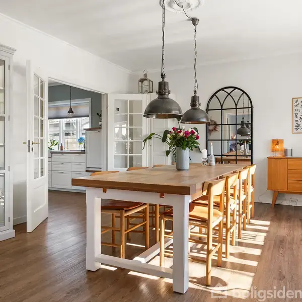 Spisebord af træ med seks stole; en vase med blomster i midten. Rummet er lyst med glasvitrine til venstre og en skænk til højre, hængende lamper over bordet.
