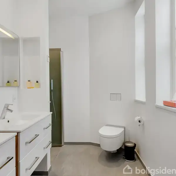 Hvidt badeværelse med vask og skuffer til venstre, toilet til højre under et vindue. Bruseniche med glasdør bagerst. Enkel og moderne indretning med lyse flader.