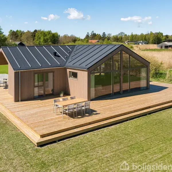 Et moderne, træbeklædt hus står stille på en stor træterrasse. Det har store, glaspartier og sort tag. Omkring er en grøn græsplæne og træer, under en klar blå himmel.