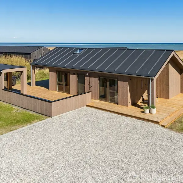 Træhus står stille på grusbelagt grund med havudsigt i baggrunden og en grøn græsplæne omkring. Terrassen er integreret i husets design.