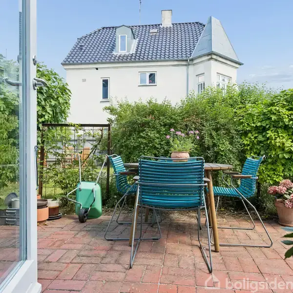 Fem blå stole står omkring et rundbord på en terrasse med blomsterkrukker. En havevogn står ved siden, omgivet af grønne buske og en hvid bygning i baggrunden.