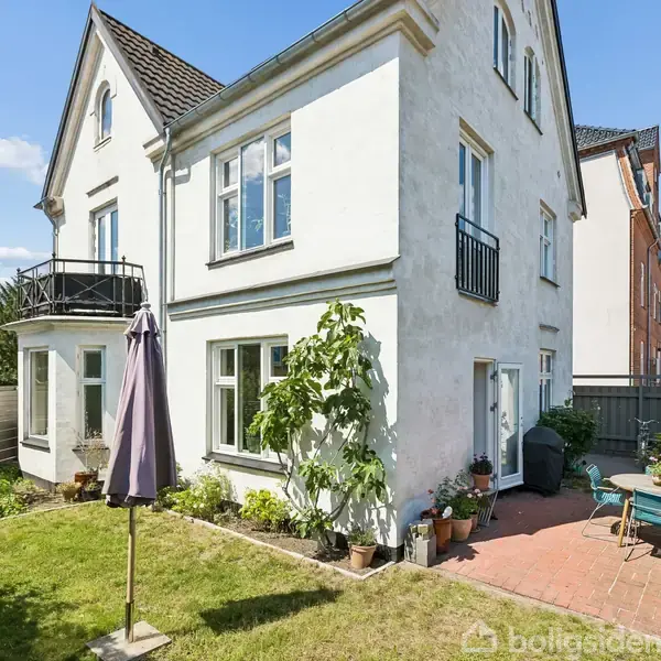 Et hvidt to-etagers hus står stille i en velplejet have, omkranset af græs, planter og en terrasse med havemøbler og parasol. Solrigt vejr og blå himmel omgiver stedet.