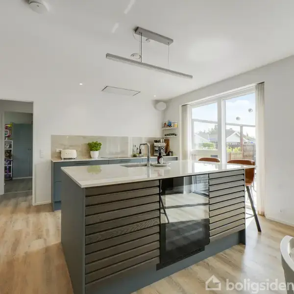Køkken med stor ø med indbygget ovn og kogeplade, omkranset af moderne trælaminatgulv. Lyse omgivelser med spiseområde, store vinduer og udsigt til terrasse. Peberkværn og plante pynter på bordet.