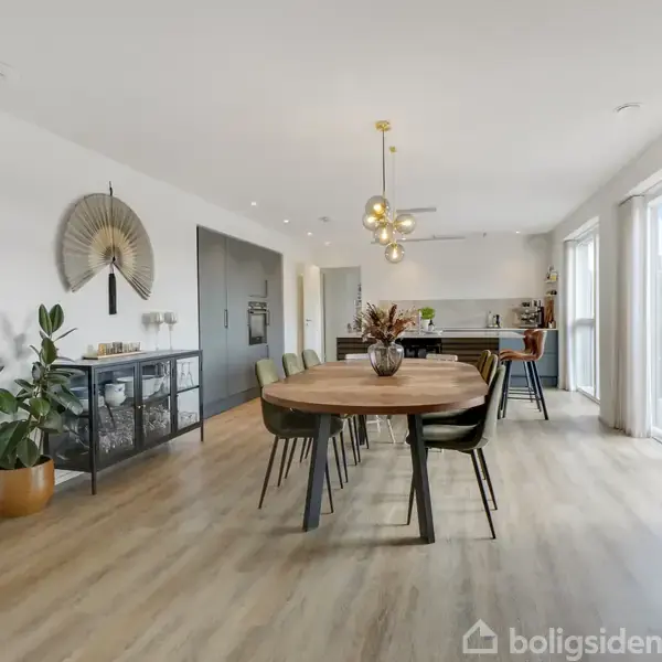 Spisebord med seks stole står i et lyst, moderne opholdsrum. Et køkken ses i baggrunden, og der er store vinduer til højre, som lukker naturligt lys ind.