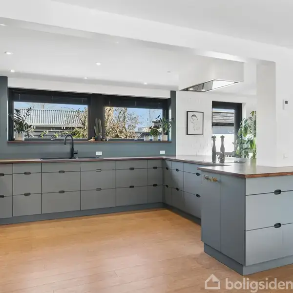 Moderne køkken med grå skabe og træbordplade. Køkkenet har store vinduer der giver naturligt lys. Planter og kunstværker indrammer rummet. Åben forbindelse til stuen ses i baggrunden.
