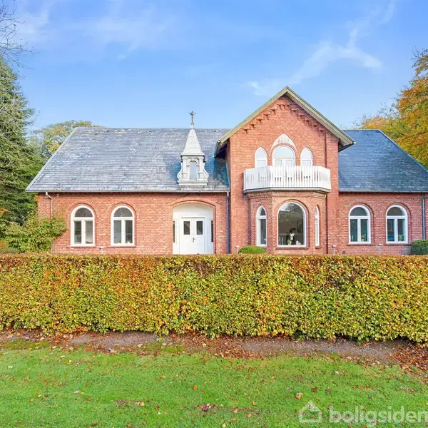 Et rødt murstenshus står stille bag en hæk, omgivet af grønne træer. Huset har en unik arkitektur med buede vinduer og en balkon, i en rolig og naturskøn have.