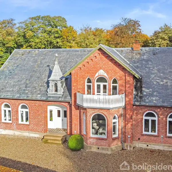 Rødt murstenshus med sort skifertag står stille omgivet af grønne buske og træer i efterårsfarver. En lille balkon pryder facaden over indgangsdøren og buede vinduer.