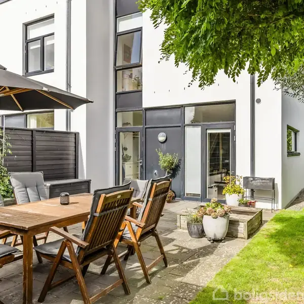 Terrassemøbler står stille under en parasol på en flisebelagt gårdhave. En moderne hvid bygningsfacade med store vinduer udgør baggrunden, omgivet af grønne planter og træer.