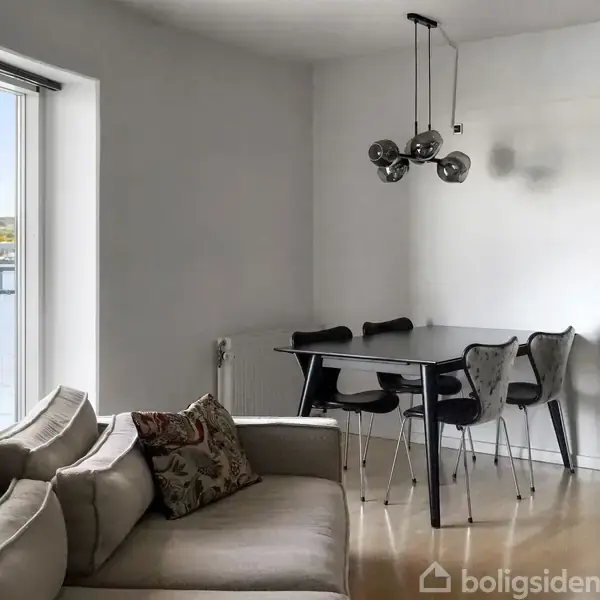 Sofa med puder står i stue ved siden af store vinduer med udsigt over vandet. Et spisebord med fire stole er placeret nær en moderne lampe.