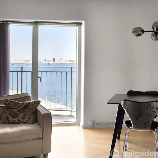 En sofa står foran en lysbilleddør, der åbner ud mod en balkon med havudsigt. Ved siden af står et spisebord med stole; en moderne lampe hænger over bordet.