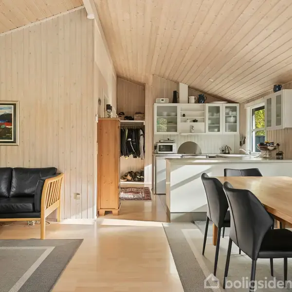 Sort lædersofa står foran et træbeklædt vægpanelmaleri. Spisebordet med sorte stole er ved siden af et åbent køkken med hvide skabe, der får naturligt lys ind fra vinduet.