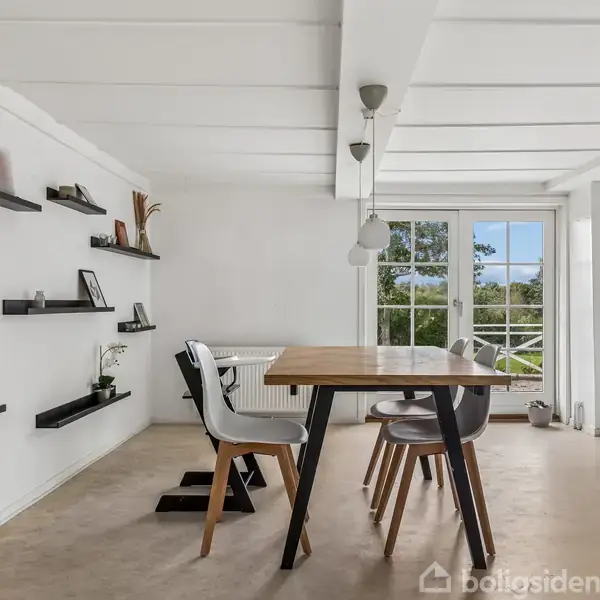 Et spisebord med stole står i et lyst rum med hvidt loft. Flydende hylder på venstre væg viser dekoration. Store vinduer i baggrunden leder ud til en balkon med udsigt.