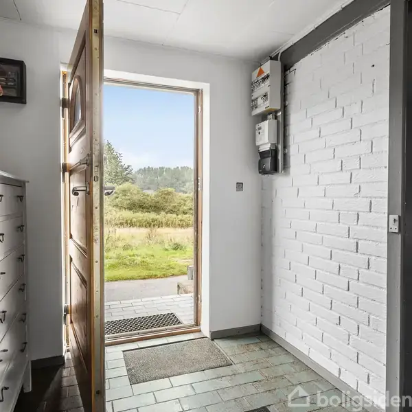 Dør står åben, lys strømmer ind fra have med græs og træer. Hvid kommode med vase og blomster til venstre, og hvide murstensvægge omkranser rummet.