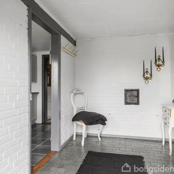 Indgangsparti med en hvid stol og sort tæppe på gråt flisegulv. Væghængt dekoration og lille hvidt bord med spejl i baggrunden. Hvide vægge.