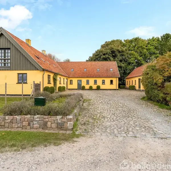 Gul, traditionel gård med rødt tegltag står for enden af en brostensbelagt indkørsel. Grønt buskads omkranser ejendommen, og træer danner en skovet baggrund under en skyet blå himmel.