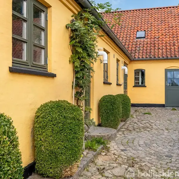Gult murstenshus med sorte vinduer, omkranser en brostensbelagt gårdsplads med klippede buske. Rødt tegltag og grøn dør tilføjer karakter. Skyfri blå himmel i baggrunden.