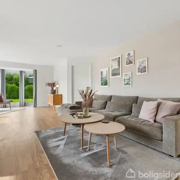En grå sofa står på et tæppe med runde borde foran, mens et gulvlampe og billeder hænger på væggen. Stuen har store vinduer med udsigt til en grøn have.