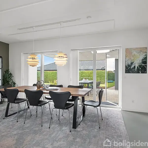 Et indendørs spiseområde med et stort træbord og seks sorte stole. To pendellamper hænger over bordet. Glasdøre i baggrunden åbner ud til en gårdhave med grønne områder.