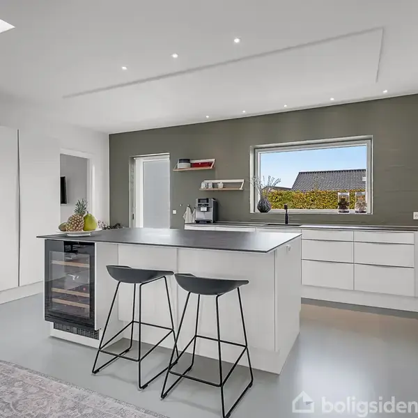 Et moderne køkken med en stor ø i midten, to barstole, indbygget vinkøleskab og hvide skabe. Store vinduer giver udsigt til en have.