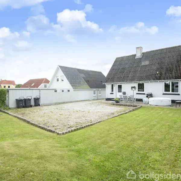 Hus med hvid facade og sort tag står i en grøn have med græsplæne og flisebelægning. Der er terrassemøbler foran huset og en hæk i baggrunden.