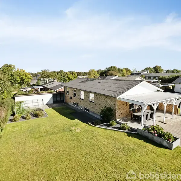 Et hus med murstensfacade ligger i en grøn have. Huset har en overdækket terrasse og er omgivet af en græsplæne og buske. I baggrunden ses andre boliger.