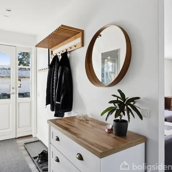Entrémøbel med plante og spejl står stille ved væggen. Jakker hænger på knagerække over en skohylde ved siden af en hoveddør. Stue ses i baggrunden.