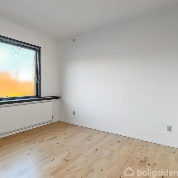 Et tomt værelse med lyst trægulv har en stor vindue til venstre, som lukker naturligt lys ind. Væggene er hvide, og der er en radiator under vinduet.